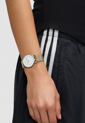 Goldfarbenes Uhrarmband aus Mesh mit rundem weißem Zifferblatt, minimalistischen Stundenmarkierungen und schlankem Profil, getragen an einem Handgelenk mit schwarzen Sporthosen.