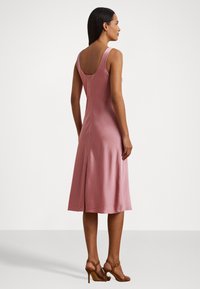 Vestido de satin rosa sem mangas, com uma parte de trás em forma de cavalo, corte em A e comprimento até aos joelhos. Textura suave com um ligeiro brilho, combinado com sapatos de salto castanhos.