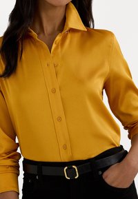 Camisa amarela de seda abotoada com colarinho, apresentando mangas longas e um cinto preto com fivela dourada. Textura suave e corte ajustado.