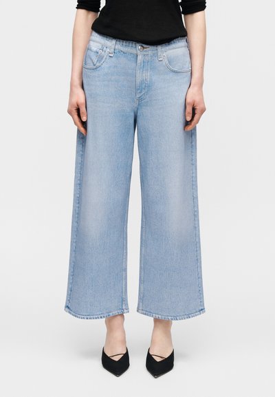 Άτομο που φοράει ανοιχτό μπλε cropped τζιν με φαρδιά μπατζάκια, μαύρο μακρυμάνικο τοπ και μαύρα μυτερά παπούτσια, στέκεται μπροστά σε απλό φόντο.