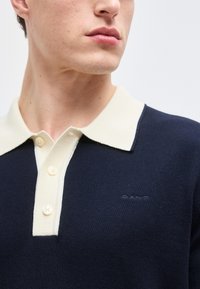 Gros plan sur un homme portant un polo en maille bleu marine avec un col crème et une patte de boutonnage, montrant une partie de son menton et de son cou.