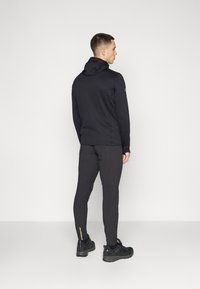 Angeschnittener schwarzer Hoodie mit strukturiertem Material, kombiniert mit schmal geschnittenen schwarzen Hosen mit reflektierenden Akzenten und schwarzen Sportschuhen.