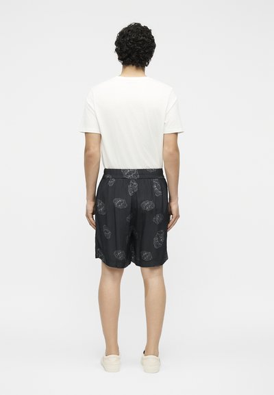 Άνδρας που στέκεται με πλάτη, φοράει λευκό κοντομάνικο πουκάμισο, μαύρα σορτς με σχέδια και λευκά αθλητικά πάνω σε λευκό φόντο.