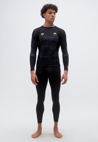 Άνδρας που στέκεται ξυπόλητος, φοράει μαύρο μοτίβο μπλουζάκι μακρυμάνικο και κολάν με χρυσά λογότυπα ζώων στο στήθος και τον καρπό, ουδέτερο φόντο.