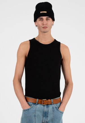 Ung mand iført sort ærmeløs skjorte, sort strikket hue, lyseblå jeans og brun bælte med hænderne i lommerne, vendt fremad.