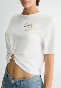 T-shirt à manches courtes blanche avec un ourlet noué, présentant un imprimé doré du logo "LIU.JO" et "EST. 1995", conçu en coton doux.
