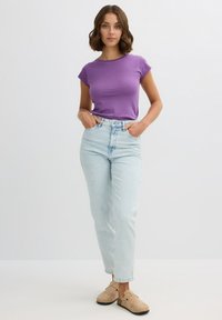 Camiseta de manga corta morada combinada con jeans de tiro alto azul claro. La modelo lleva unas zapatillas de terciopelo beige y está de pie con las manos en los bolsillos.