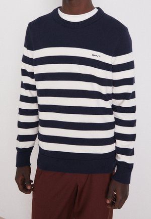 Dunkelhäutige Person trägt einen marineblau-weiß horizontal gestreiften Pullover mit "GANT"-Logo und dunkelrote Hose, steht vor schlichter Hintergrund.