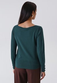 Vrouw met lang, donker haar die gouden oorringen draagt, een donkergroene trui met lange mouwen en een broek in bruin, met haar rug naar de camera gekeerd tegen een effen achtergrond.