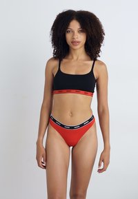 Svart bralette med rød stripe med "HUGO"-tekst, kombinert med rød truse med svart midjebånd. Glatt stoff, minimalistisk design.