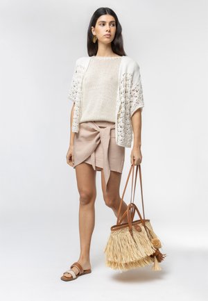 Woman wearing loose white knit top and beige wrap skirt, holding large straw tote bag with leather handles, standing on white background.