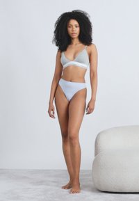 Γκρι σουτιέν με λευκή ταινία και ανοιχτό γαλάζιο σλιπ με ψηλή κοπή. Το μοντέλο στέκεται σε ουδέτερο χαλί, με έναν μινιμαλιστικό μπεζ καναπέ κοντά του.