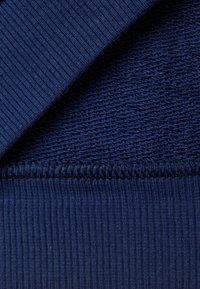 Tessuto a maglia blu navy che mostra due trame: una sezione a coste e una tessitura più liscia e annodata con cuciture visibili lungo il bordo.
