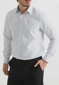 Homme portant une chemise à boutons à manches longues blanche avec de petits pois noirs et un pantalon noir, la main dans la poche, sur fond uni.