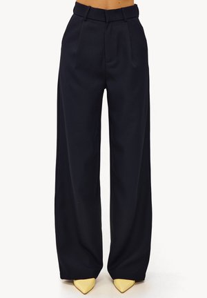 Pantalon large bleu marine en tissu lisse, avec une ceinture ajustée et des plis, associé à des escarpins jaunes à bout pointu.