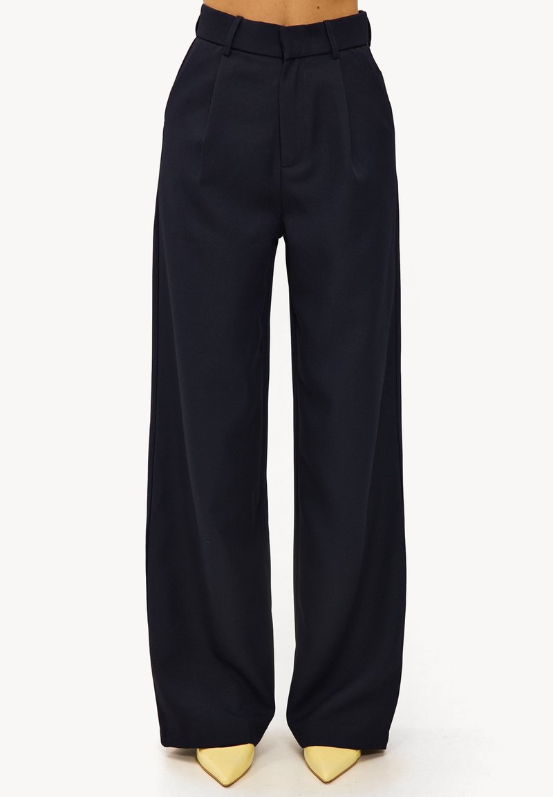 Pantalon large bleu marine en tissu lisse, avec une ceinture ajustée et des plis, associé à des escarpins jaunes à bout pointu.