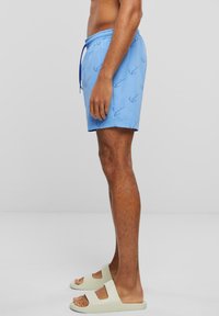 Shorts de bain bleus avec une taille à cordon de serrage et un motif subtil. Portés avec des sandales de couleur neutre. Tissu lisse et léger.