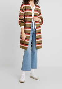 Cárdigan de punto en rayas marrón, rosa y navy, con mangas largas y abertura en el frente. Combinado con vaqueros acampanados de color azul claro y zapatos blancos.