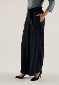 Black wide-leg pants with a textured finish, featuring a belted waist and side pockets, paired with black pointed-toe heels.