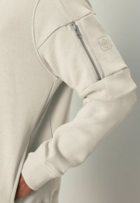 Sudadera de color beige claro con puños acanalados y un bolsillo con cremallera en la parte superior de la manga. Presenta una textura suave y costuras redondeadas en los hombros.