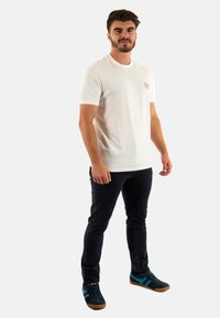 T-shirt bianca in cotone con maniche corte, caratterizzata da un piccolo logo rotondo. Combinata con jeans in denim scuro e sneakers nere con dettagli blu.