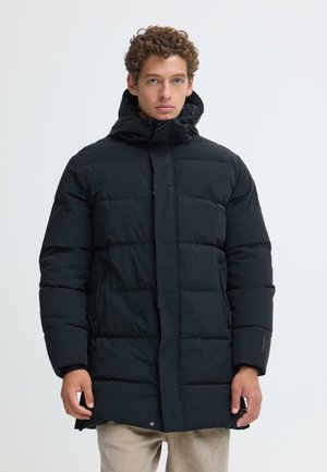 Homme aux cheveux bouclés portant une longue doudoune noire à capuche et un pantalon beige, debout devant un fond blanc uni.