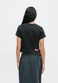 Γυναίκα με μακριά μαύρα μαλλιά φοράει ένα μαύρο κοντό μπλουζάκι και σκούρα φούστα, στέκεται με την πλάτη προς την κάμερα μπροστά σε απλή φόντο.