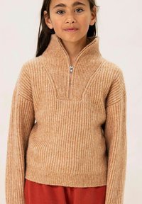 Beige gerippter Pullover mit hohem Kragen und halbem Reißverschluss. Weiche Textur, lockere Passform und lange Ärmel. Getragen mit hellroten Hosen.
