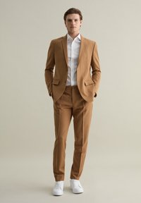 Traje color arena con corte ajustado, chaqueta de un solo botón, pantalones a juego y camisa blanca con cuello. Combinado con zapatillas blancas.