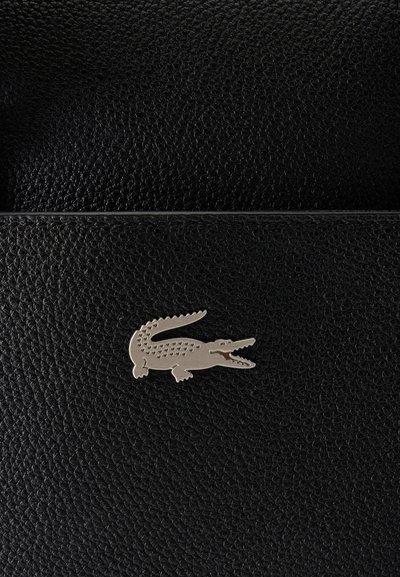 Surface en cuir noir texturé avec un petit logo métallique de crocodile au centre, ressemblant à un emblème de marque.