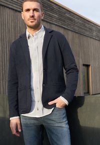 Man in een marineblazer, wit overhemd en blauwe spijkerbroek staat buiten tegen een houten muur met één hand in zijn zak.