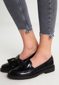Mocasines de charol negro con detalle de borlas, puntera redondeada y suela de goma texturizada baja, combinados con jeans ajustados gris deshilachados.