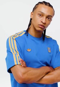 Mladić s pletenicama odjeven u plavu sportsku majicu sa žutim prugama, prekriženih ruku, gleda izravno u kameru.