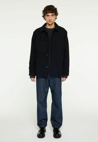 Svart ullblandad jacka med knäppning, oversized passform och spetsig krage, tillsammans med mörkblå denimbyxor och svarta skor.