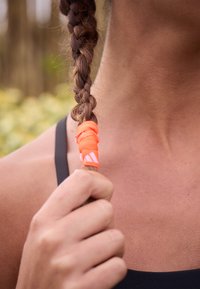 Ein geflochtener, dunkelbrauner Haarschopf, gehalten von einem leuchtend orangefarbenen Gummiband. Der Hautton ist gebräunt; der Hintergrund zeigt unscharfes Grün.