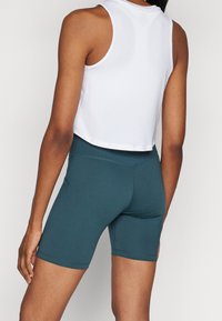 Top corto bianco abbinato a pantaloncini da ciclismo verde petrolio a vita alta, con tessuto liscio e design aderente, che mette in risalto la vista posteriore.