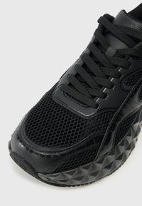 Schwarzer Sportschuh mit einem Mesh-Obermaterial, strukturierten Akzenten und einer dicken, dreieckig gemusterten Sohle. Schwarze Schnürsenkel und verstärkter Zehenbereich.