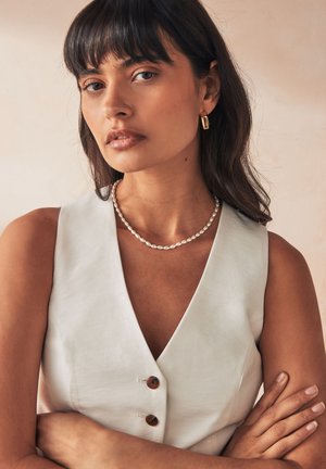 Frau mit mittellangen dunklen Haaren, die eine ärmellose weiße Weste, eine Perlenkette, goldene rechteckige Ohrringe trägt und die Arme verschränkt hat.