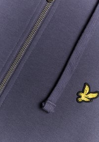 Lila Kapuzenjacke mit Reißverschluss, goldfarbenem Reißverschluss und gesticktem gelbem Vogel-Logo auf der linken Brust, gefertigt aus einem glatten Stoff mit strukturiertem Kapuzenfutter.