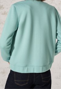 Osoba oblečená v světle modré mikině a tmavě modrých džínách, zobrazená zezadu na neutrálním texturovaném pozadí.