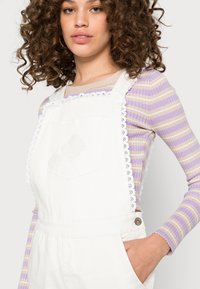 Macacão branco de algodão com acabamentos em renda, apresentando um bolso e detalhes de botões. Por baixo, uma blusa de mangas longas listrada em lavanda e bege.