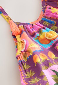 Primer plano de un vestido púrpura colorido con patrones de frutas tropicales y hojas, con un tassel rosa en una de las tiras del hombro.