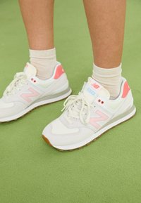 Baskets blanches New Balance avec accents roses, design à lacets, dessus texturé et semelle en caoutchouc marron. Portées avec des chaussettes montantes beige clair.