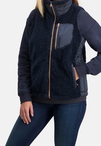 Marinblå väst som kombinerar fluffig tyg och släta, quiltade sektioner. Har dragkedja, sidfickor och hög krage med en hållbar struktur.