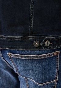 Denim jacket with dark blue fabric, contrasting orange stitching, two metal buttons at the waist, and a back pocket with a label.