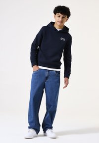 Sudadera azul marino con bolsillo frontal y logo blanco, combinada con jeans de mezclilla azul de corte holgado y zapatillas blancas sobre un fondo liso.