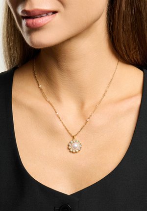 Kvinde iført guldkæde med vedhæng formet som en rund perle og opalblomst på en sort top.