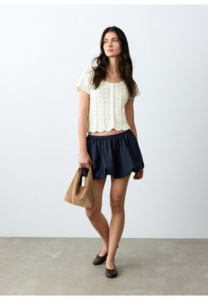 Žena s dlhými tmavými vlasmi, oblečená v bielom pletenom tričku s krátkym rukávom a námornícky modrej sukni, drží hnedú kabelku a stojí na šedom podlahe.