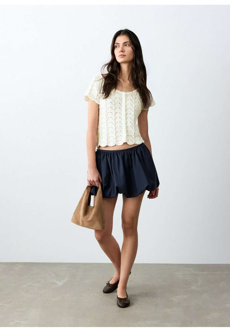 Ženska z dolgimi temnimi lasmi, oblečena v belo pleteno kratkoročno majico, mornarsko modro krilo, drži rjavo ročno torbico, stoji na sivi tla.