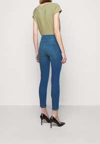 Jeans ajustados de color azul con cintura alta y bolsillos traseros, combinados con una blusa de manga corta en verde oliva y zapatos de tacón negro.